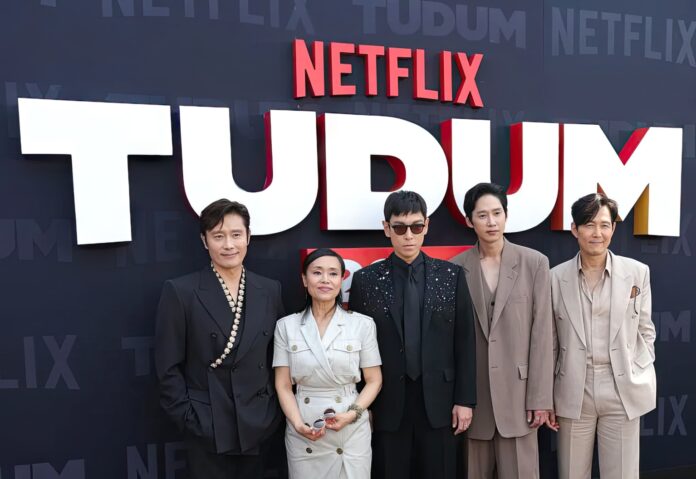 Large crowd of Netflix fans attending Tudum event, waving hands and holding merchandise.