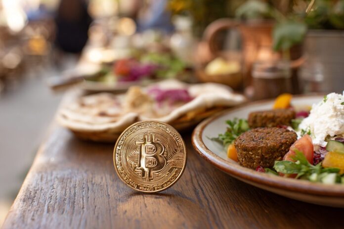 Gold Bitcoin coin on a wooden restaurant table beside a plate of Middle Eastern food