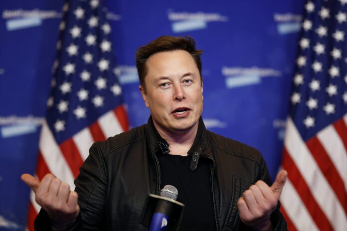 Elon Musk speaking at a press event, gesturing energetically with a backdrop of U.S. flags and tech logos.