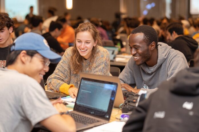 A diverse group of college students sit around a table, working together with laptops and AI-powered educational tools on display.