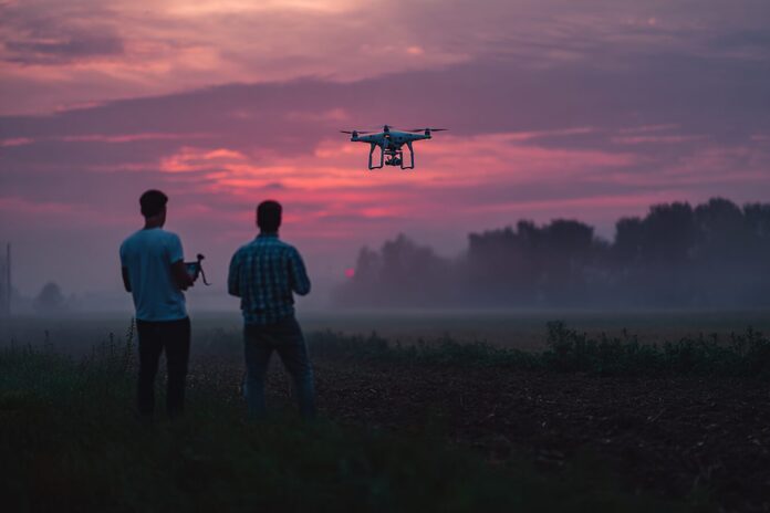 Two men operating remote controls and observing a drone in flight over a field at dusk