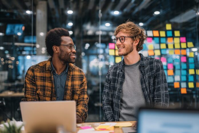 Two diverse engineers discussing project details in a modern workspace with post-it notes and laptops.