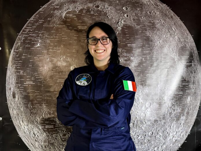 Benedetta Facini in a NASA flight suit, smiling and standing in front of a mission emblem backdrop.