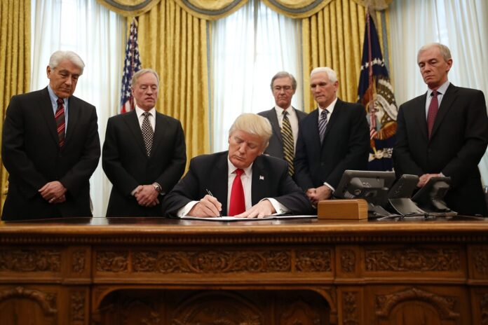 President Trump signs executive order at a desk, surrounded by advisors
