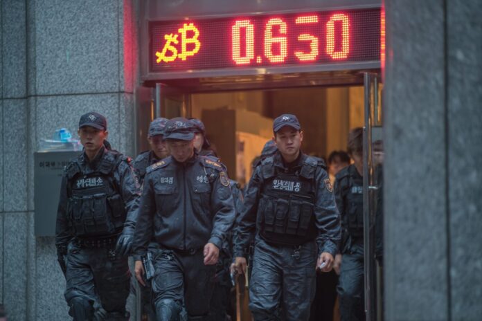 South Korean police leading handcuffed cybercriminal suspects from a building, with a digital display of cryptocurrencies in the background.