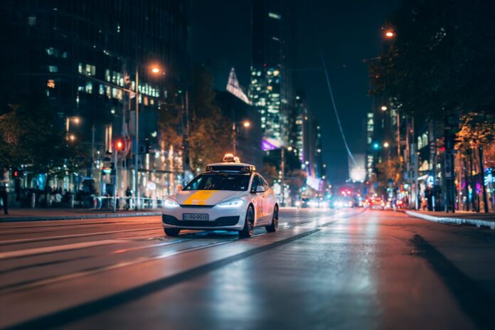 Robotaxi vehicle navigating an urban city street at night, illuminated by city lights and other cars.