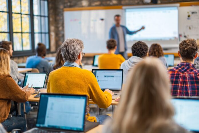 A group of diverse adults in a bright classroom using laptops while an instructor points at an AI-powered presentation on a screen.