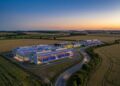 Aerial view of a modern data center campus near Paris at dusk with glowing server lights and surrounding green fields under a clear sky.